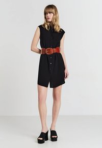 Abito nero con maniche corte, frontale con bottoni e vita cintrata accentuata da una cintura in pelle marrone. Scarpe nere con zeppa.