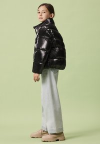 Giacca imbottita nera lucida con collo alto. Abbinata a jeans azzurri a gamba larga e scarpe beige con suola spessa. Look casual nel complesso.