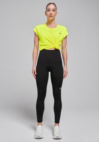 Haut à manches courtes jaune fluo noué à la taille, associé à un legging noir taille haute et des baskets blanches.
