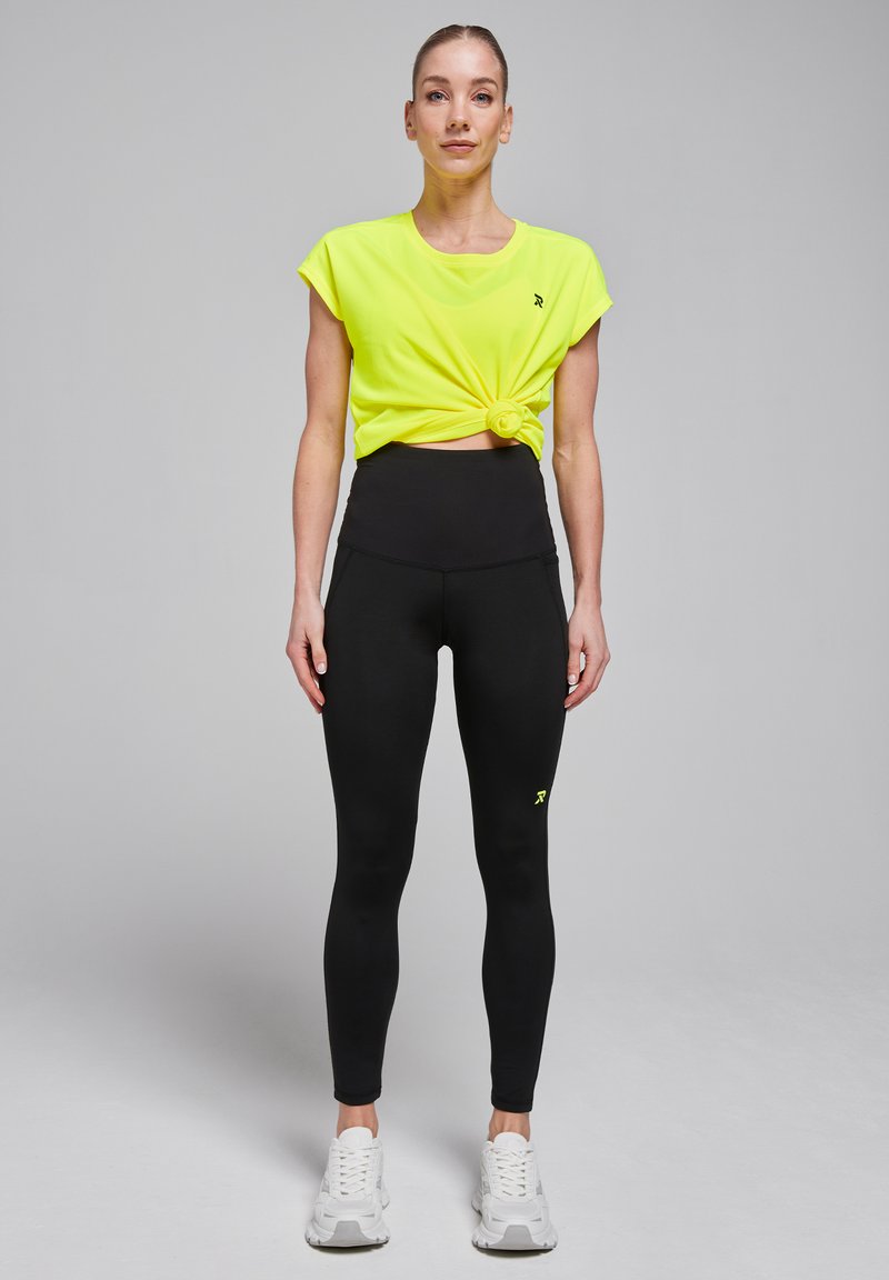 Haut à manches courtes jaune fluo noué à la taille, associé à un legging noir taille haute et des baskets blanches.