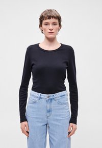 Μαύρη μπλούζα με μακριά μανίκια και στρογγυλή λαιμόκοψη, συνδυασμένη με ψηλόμεσα ανοιχτόχρωμα τζιν. Λεπτός ύφασμα και εφαρμοστό στυλ.