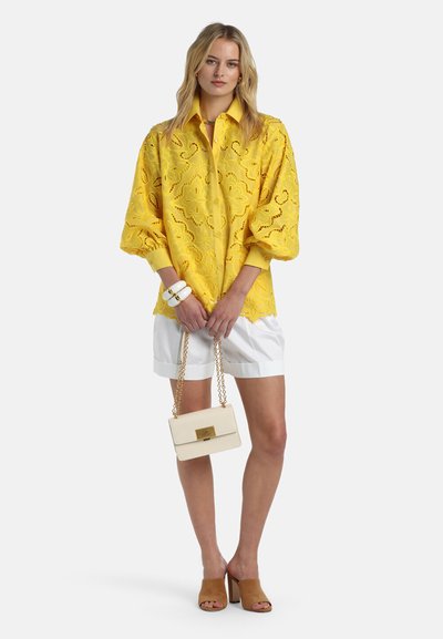 Femme blonde portant un chemisier jaune en broderie anglaise à manches ballon, un short blanc, des mules à talons couleur beige, un bracelet blanc et tenant un petit sac à main crème.