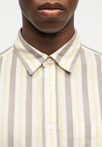 Κοντινό πλάνο ενός ατόμου που φοράει πουκάμισο με κουμπιά, με κάθετες ρίγες σε μπεζ, λευκό και κίτρινο, εμφανίζοντας τον γιακά και το πάνω μέρος του στήθους.