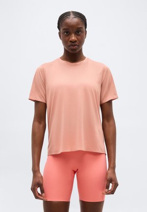 Γυναίκα με πλεξούδες φοράει φαρδιά ροζ κοντομάνικη μπλούζα και εφαρμοστά κοραλλί ποδηλατικά σορτσάκια, στέκεται μπροστά σε απλό ανοιχτό φωτισμένο φόντο.