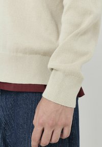 Pull en maille beige avec des poignets côtelés, superposé sur une chemise bordeaux, associé à un pantalon bleu marine à rayures. Accent sur les textures et les couleurs.