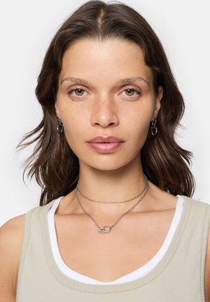 Zilveren gelaagde kettingen met een knoopdetail, gedragen met een lichtbeige tanktop met een witte rand. Gladde, glanzende huid met sproeten.