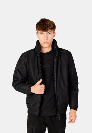Young man wearing a black zip-up jacket over a black Calvin Klein shirt, standing against a plain light gray background.