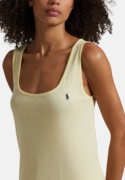 Femme aux cheveux bouclés portant un haut sans manches jaune pâle avec un petit logo sombre de cheval et cavalier sur la poitrine.