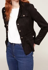 Svart mockajacka med en figursydd design, tryckknappar och två framfickor. Bärs över en vit topp med blåjeans.