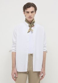 Giovane uomo vestito con una camicia bianca a bottoni sopra una canottiera bianca, pantaloni beige e una sciarpa a pois beige, in piedi contro uno sfondo neutro.