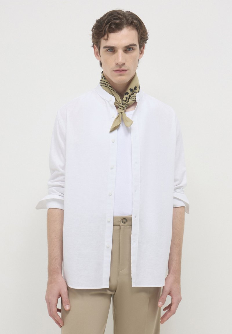 Giovane uomo vestito con una camicia bianca a bottoni sopra una canottiera bianca, pantaloni beige e una sciarpa a pois beige, in piedi contro uno sfondo neutro.