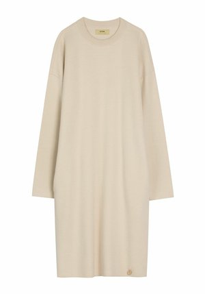 Beżowa, dzianinowa sukienka na długi rękaw, sięgająca do kolan, z okrągłym dekoltem i małym, okrągłym złotym wisiorkiem przy dolnym brzegu.