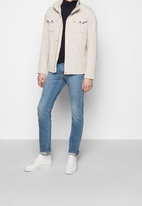 Giacca beige con due tasche sul petto e bottoni a pressione, indossata sopra un top scuro. Jeans azzurri chiari e sneakers bianche completano l'outfit.