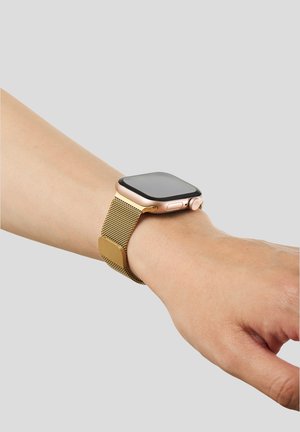 Smartklokke med gullflettet rem på venstre håndledd mot ensfarget lys grå bakgrunn.