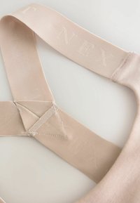 Beige tygrem med präglade bokstäver "NEXT." Platt struktur, triangel-form där remmarna korsar varandra, sydda detaljer längs kanterna.