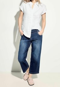 Camicia bianca in lino a maniche corte con bottoni, jeans larghi blu scuro a vestibilità rilassata e scarpe slip-on rosa chiaro con fibbie.