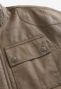 Chaqueta de cuero marrón con una textura suave, que presenta un bolsillo en el pecho con botones de presión plateados y costuras detalladas.