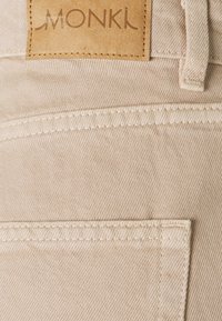 Beige denimo tkanina z blízkým záběrem, zobrazující viditelnou kapsu, detaily šití a koženou visačku s raženým nápisem "MONKĪ".