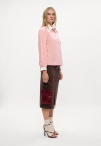 Blusa rosa a maniche lunghe con colletto bianco e dettagli decorativi, abbinata a una gonna marrone fino al ginocchio e una piccola borsa di paillettes rossa.