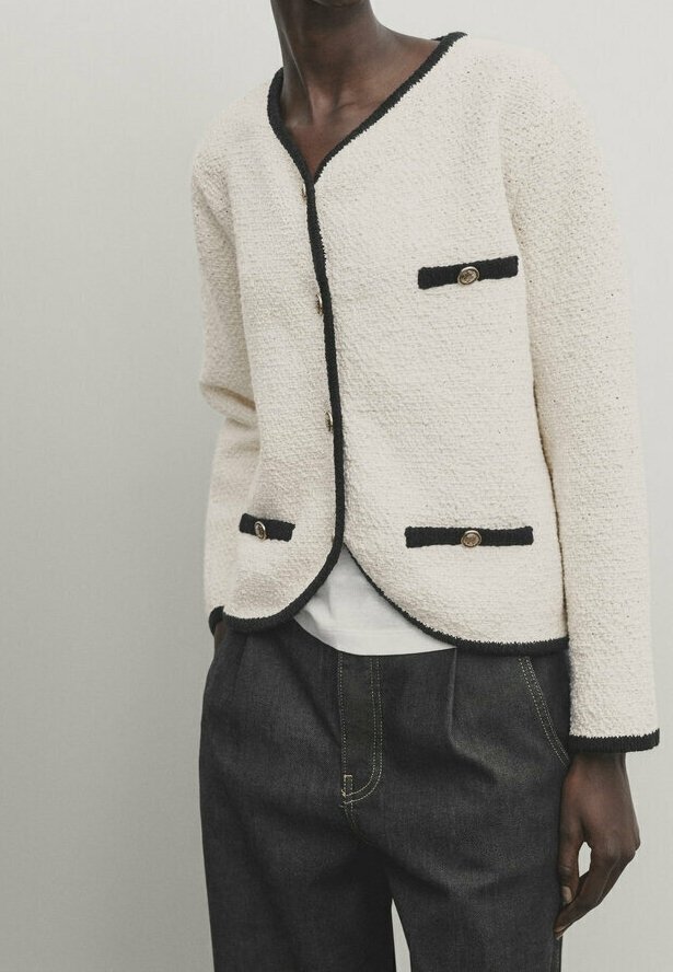 Kurtka o kremowej teksturze z czarnymi wykończeniami, z dwoma przednimi kieszeniami i złotymi guzikami, w połączeniu z ciemnoniebieskimi spodniami dżinsowymi.