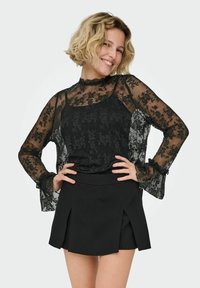 Černý krajkový top s dlouhými, zvonovými rukávy přes úzký černý tílko, doplněný černou skládací mini sukni. Celoplošný texturovaný květinový vzor.