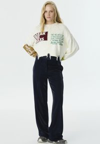 Pull blanc court avec ourlet effiloché, arborant un texte graphique coloré. Associé à un pantalon large bleu marine. Des chaussures de sport décontractées complètent le look.