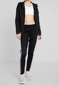 Czarna sportowa kurtka z kapturem, z teksturowanym wzorem; połączona z czarnymi zwężanymi spodniami z białymi paskami po bokach i jasnoszarymi trampkami.