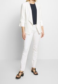 Blazer bianco con colletto a scialle, abbinato a un top blu navy, pantaloni bianchi slim-fit e sandali neri con un design a fascia.