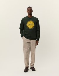 Σκούρο πράσινο φούτερ με κίτρινο κυκλικό λογότυπο, συνδυασμένο με ανοιχτό μπεζ παντελόνι καμπάνα και καφέ παπούτσια με κορδόνια σε ουδέτερο φόντο.