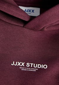 Gros plan sur un tissu bordeaux avec un texte blanc indiquant JJXX Studio, proposé par Jack & Jones, conçu à Copenhague, et une étiquette de vêtement JJXX Girl.