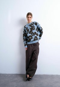 Jeune femme appuyée contre un mur blanc, portant un pull à motifs bleu et marron, un pantalon large noir et des chaussures noires pointues.