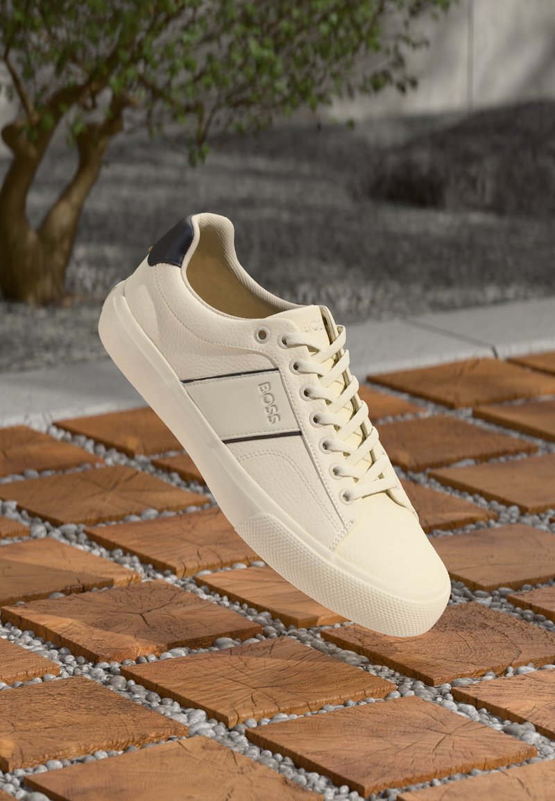 White leather sneaker with a textured finish, featuring a black heel tab and a contrasting stripe. Lace-up design with rounded toe.