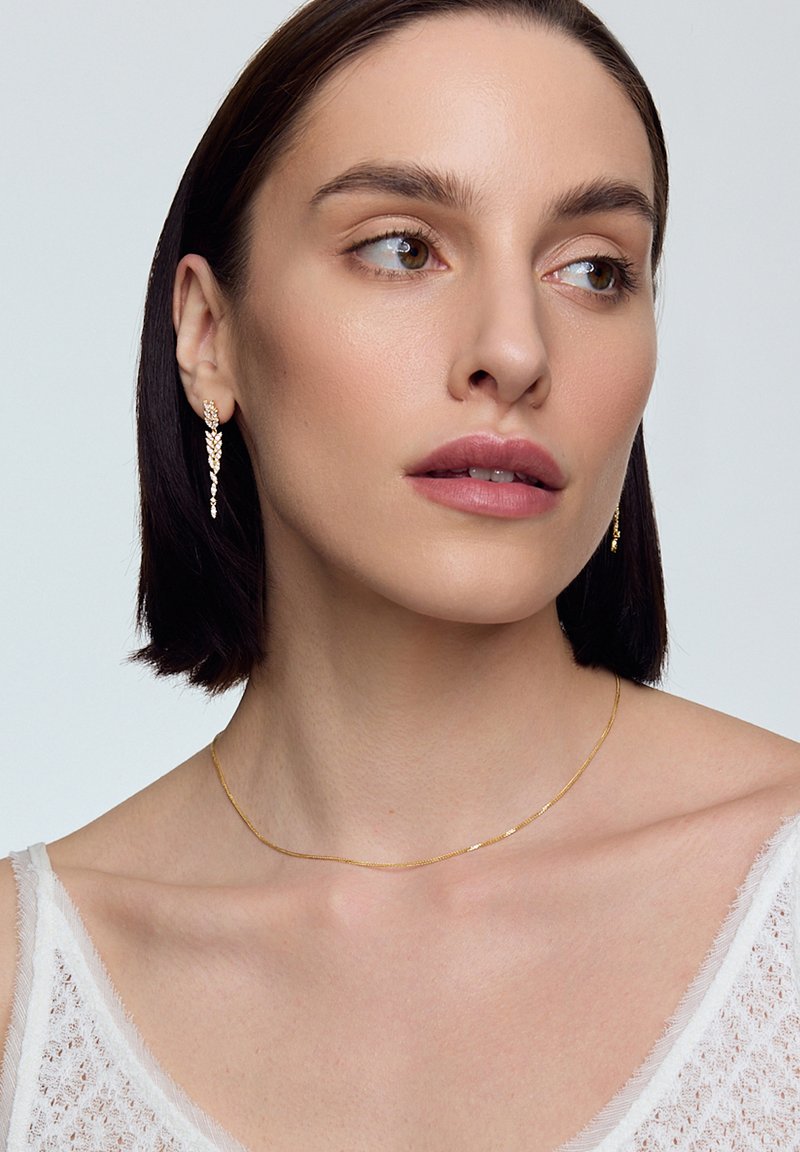 Złote kolczyki z kaskadowym łańcuchowym wzorem, w połączeniu z delikatnym złotym naszyjnikiem, noszone z białą, fakturowaną bluzką.