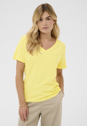 Vrouw met golvend blond haar, die gouden oorringen draagt, een gele V-hals T-shirt, beige broek en gouden armbanden, tegen een effen achtergrond.