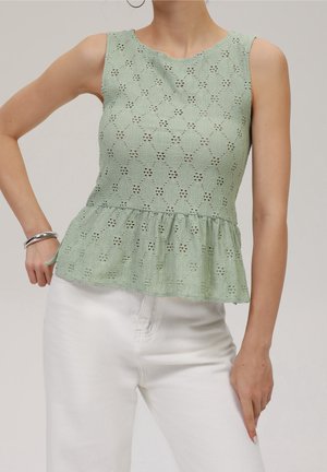Top à basque vert en dentelle sans manches porté avec un pantalon blanc, accessoirisé d’un bracelet argenté et de boucles d’oreilles créoles, sur fond uni.