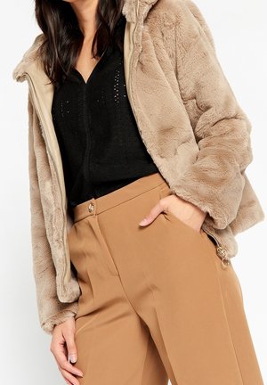 Kvinde iført en beige faux fur jakke over en sort top og camel-farvede højtaljede bukser med hånden i lommen.