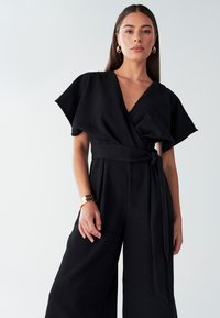 Črn kombinezon z zavitki, širokimi rokavi, izrezom v obliki črke V in pasom, ki se veže. Gladek tekstura z nabrano zasnovo in širokimi hlačnicami s sproščenim krojem.