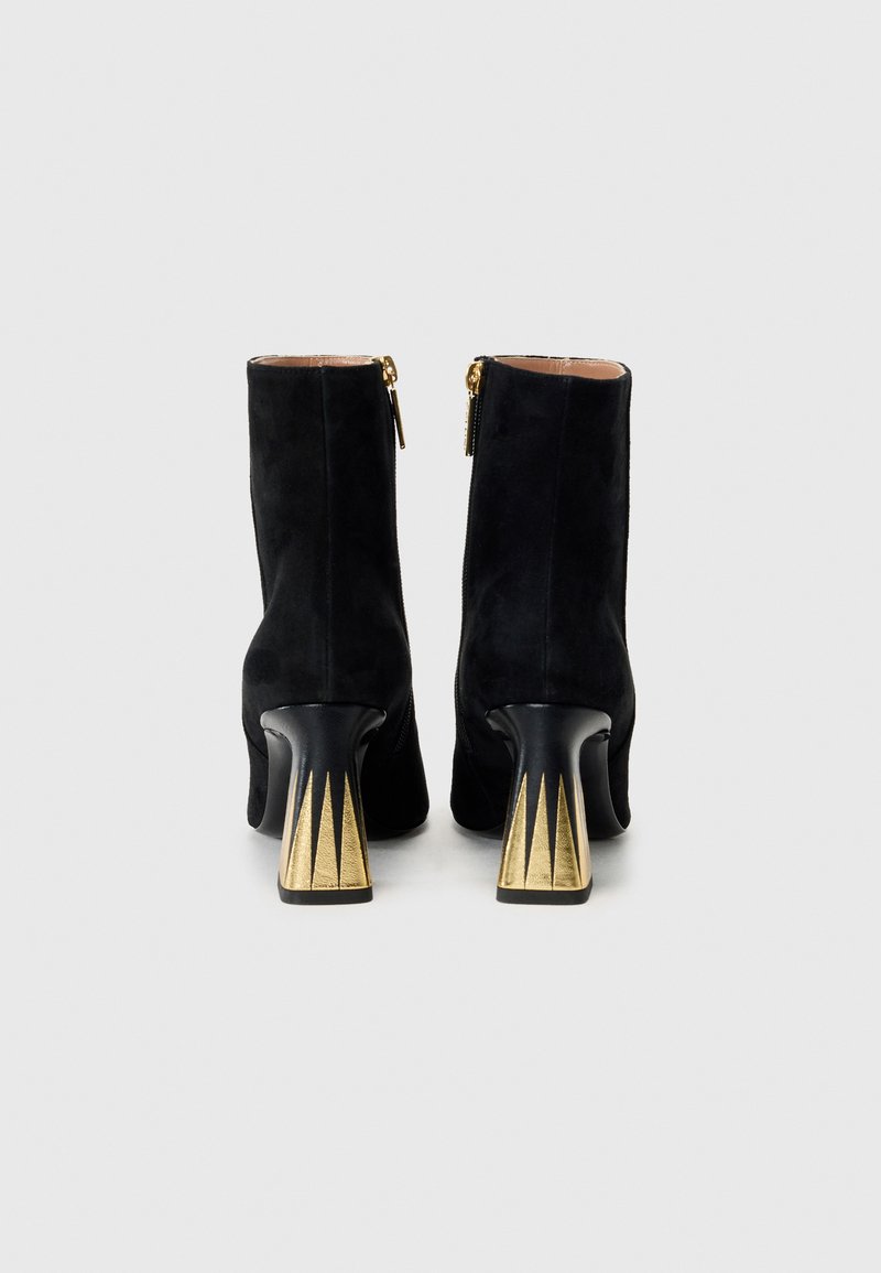 Une paire de bottines en daim noir avec une fermeture éclair dorée et des talons noirs sculptés, présentant des accents triangulaires dorés, vues de l'arrière.