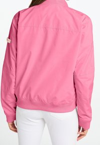 Rosa bomberjacka med ribbat krage och manschetter, med en slät textur och sidofickor. Baksidan är enhetlig utan logotyper eller dekorationer.