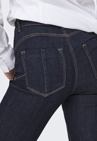 Los jeans de mezclilla azul oscuro presentan bolsillos traseros con costuras visibles, un diseño ajustado y una textura suave. Se puede ver parcialmente una camisa blanca.