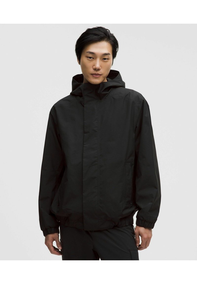 Młody mężczyzna ubrany w czarną kurtkę z kapturem, z elastycznymi mankietami i bocznymi kieszeniami, stojący na tle jednolitego jasnoszarego tła.