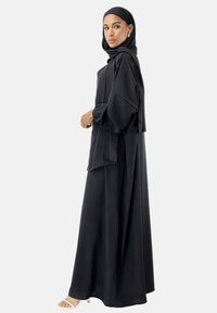 Schwarze, langärmelige Abaya mit fließender Silhouette und weichem Material. Enthält einen passenden Hijab und subtile Drapierungsdetails. Weiße Sandalen vervollständigen den Look.