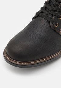 Scarpa in pelle nera con punta arrotondata, superficie testurizzata, cuciture chiare a contrasto e lacci intrecciati, che offrono un design classico.