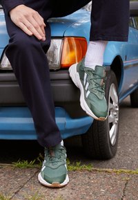 Green sneakers with white accents, textured fabric, and a gum sole. Worn with white socks and navy pants, resting on a car bumper.