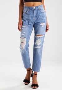 Jeans de mezclilla de tiro alto en azul claro, con parches desgastados. Corte relajado con dobladillos deshilachados y detalles de costura visibles.