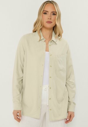 Kobieta o blond włosach, ubrana w oversize beżową koszulę na guziki, na białej bluzce i białych spodniach na jednolitym tle.