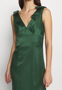 Nainen, joka on pukeutunut senza hihoja tumman vihreään satiinimekkoon, jossa on V-pääntie ja nauha-yksityiskohtia olkaimissa, seisoo valkoista taustaa vasten.