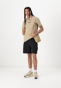 Beige tričko s krátkým rukávem s logem Timberland, černé cargo šortky, béžové a bílé tenisky a malá béžová crossover taška.