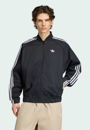Junger Mann mit lockigem Haar trägt eine schwarze Adidas Trainingsjacke mit weißen Streifen und beige Hosen, steht vor einfarbig hellem Hintergrund.