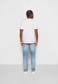 Maglietta bianca a maniche corte con bordo nero sulle maniche, abbinata a jeans blu chiaro. Fit semplice e casual, vista da dietro.